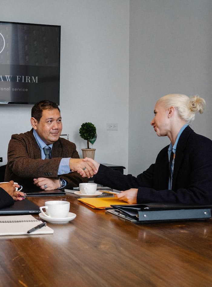 Smiling multiracial coworkers in formal clothes gathering at wooden table with cups of coffee and various folders and shaking hands while looking at each other